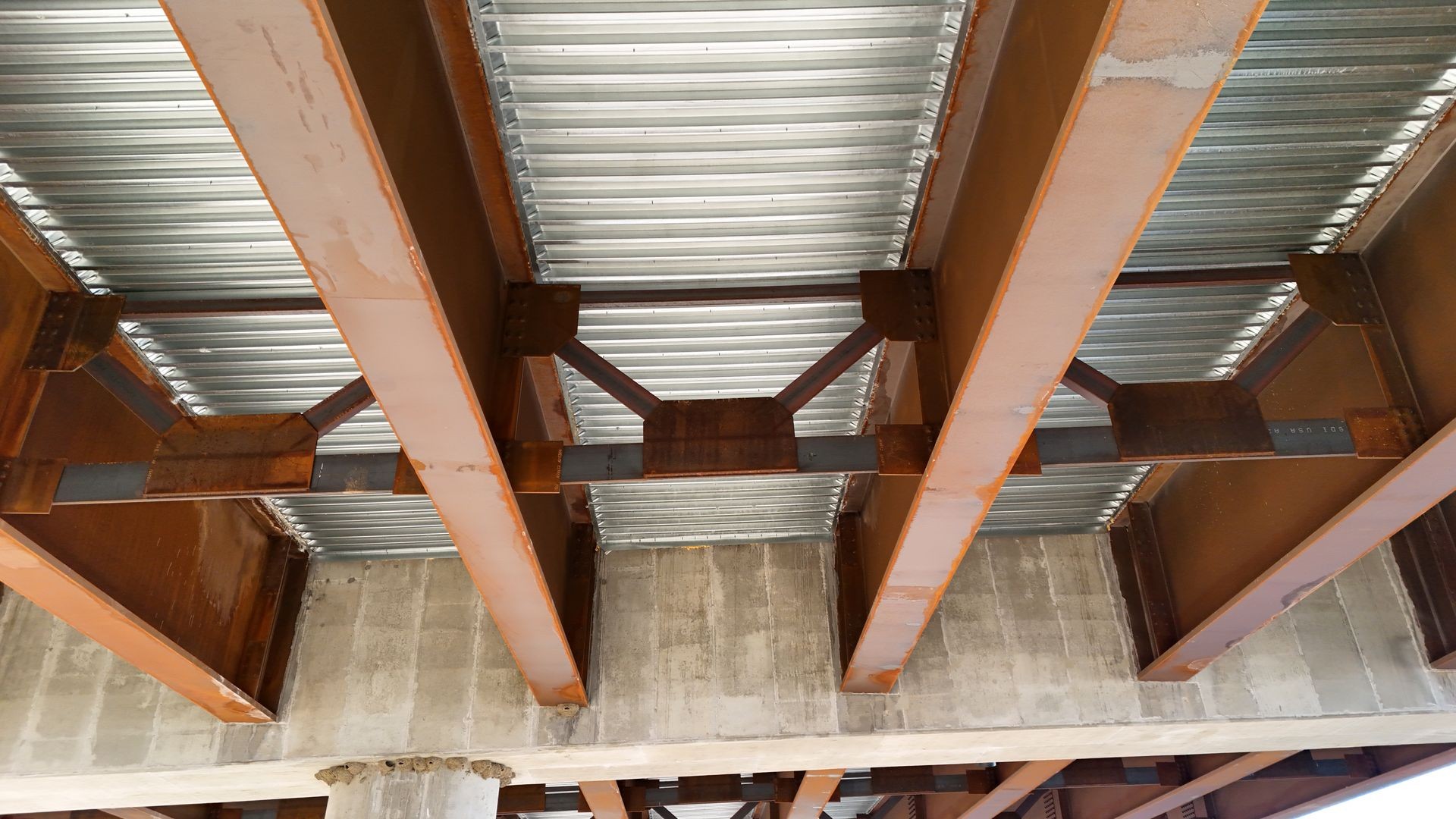 Underneath view of a metal bridge with steel support beams and concrete piers.